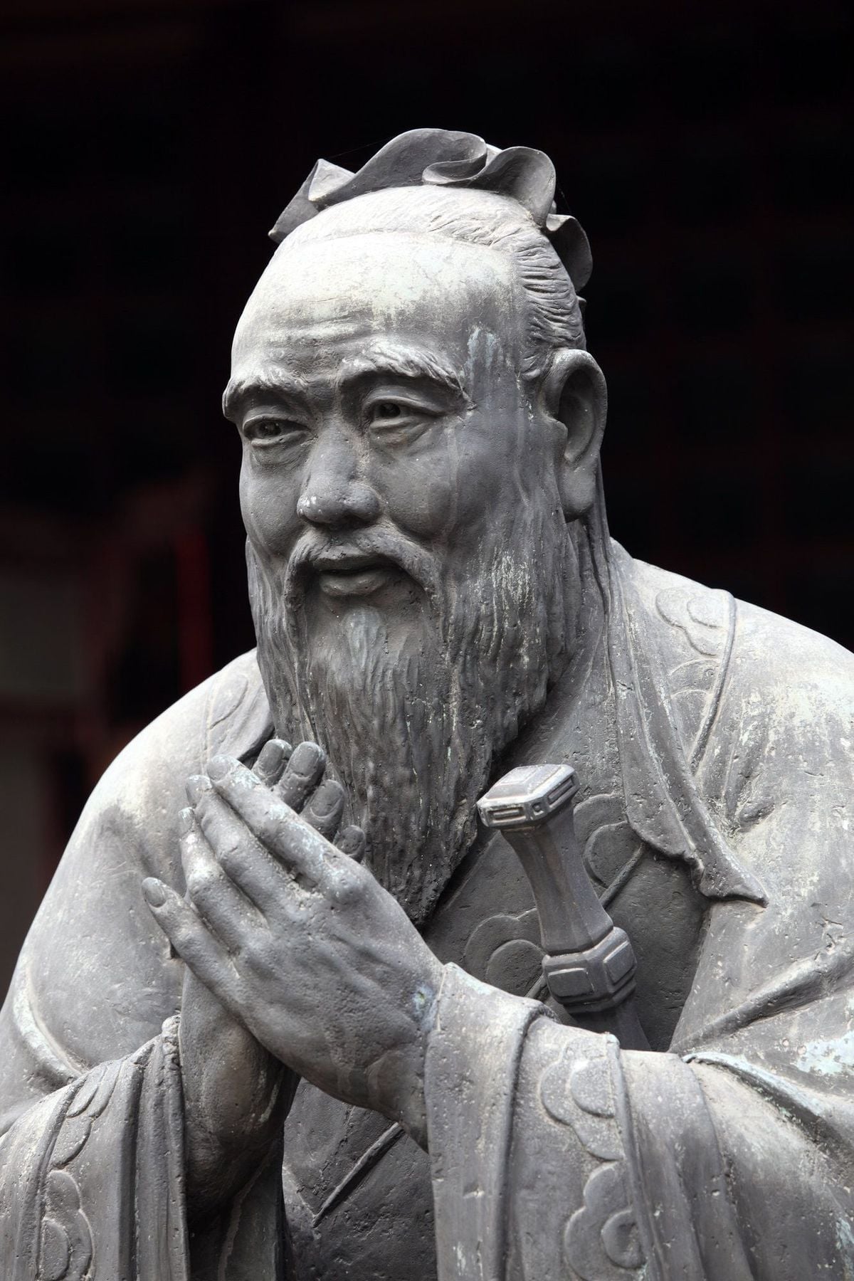A stone statue of a bearded man with hands clasped together, wearing traditional robes. The figure has a calm expression and his hair is styled in a topknot.