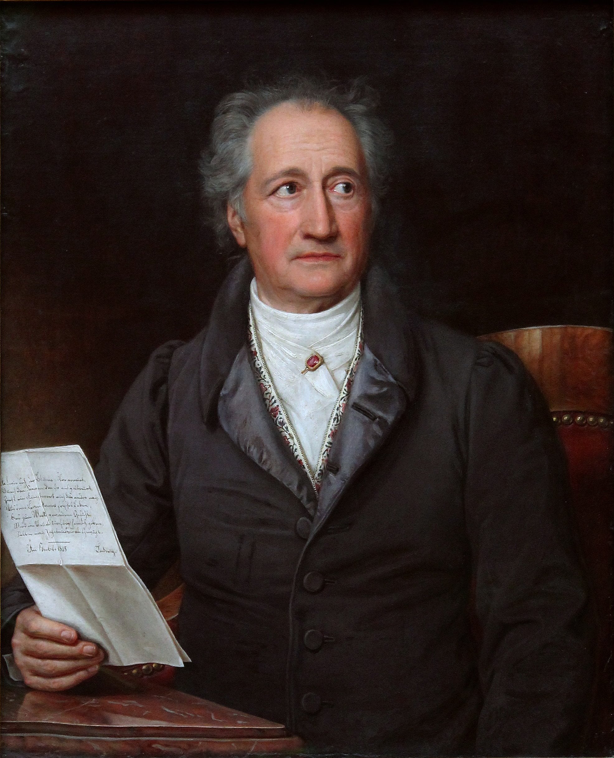 An older man with gray hair sits in a chair, wearing a dark coat and white shirt, holding a handwritten letter in one hand and looking slightly to his left against a dark background.