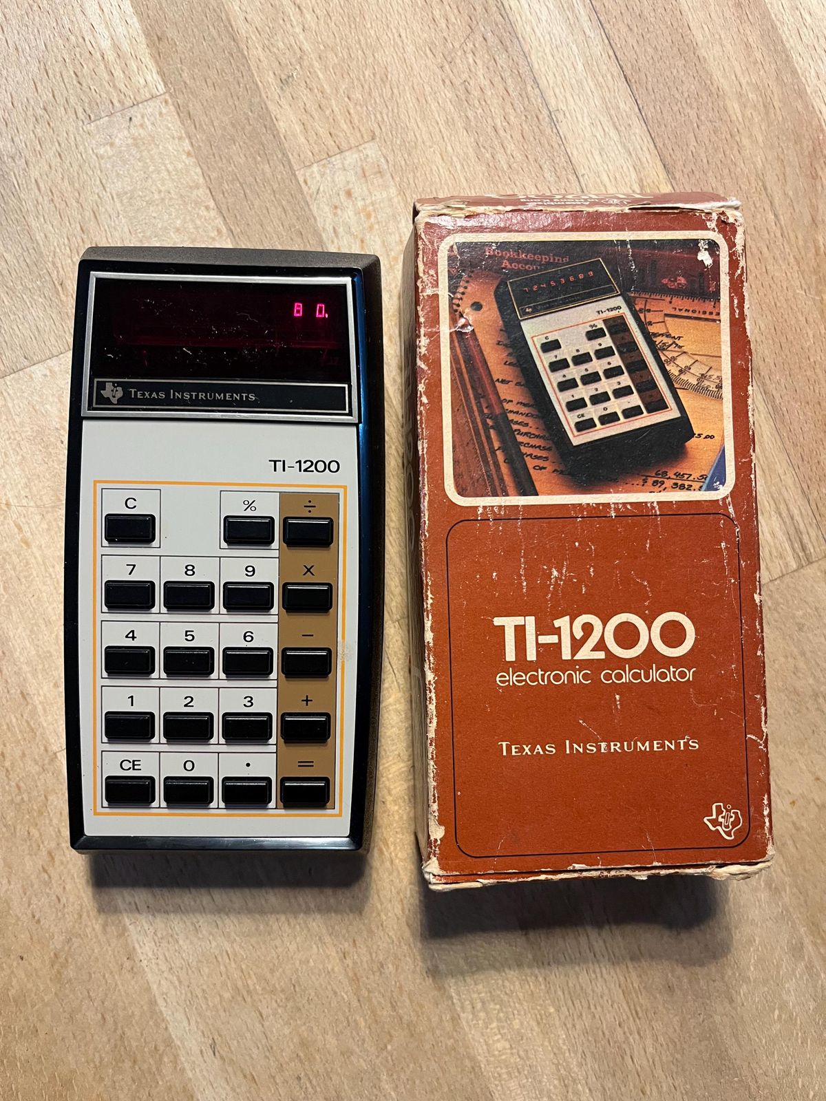 A vintage Texas Instruments TI-1200 calculator sits next to its original worn box, both on a wooden surface. The calculator’s display shows "8.0".