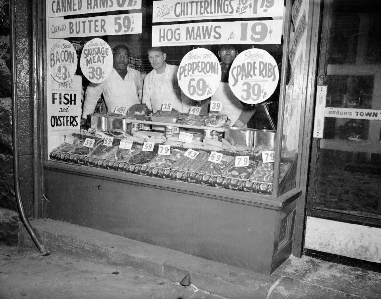 Three men stand behind a meat counter in a shop window displaying sausages, ribs, and signs showing prices for various meats like bacon, pepperoni, and spare ribs. The window is filled with handwritten price signs.