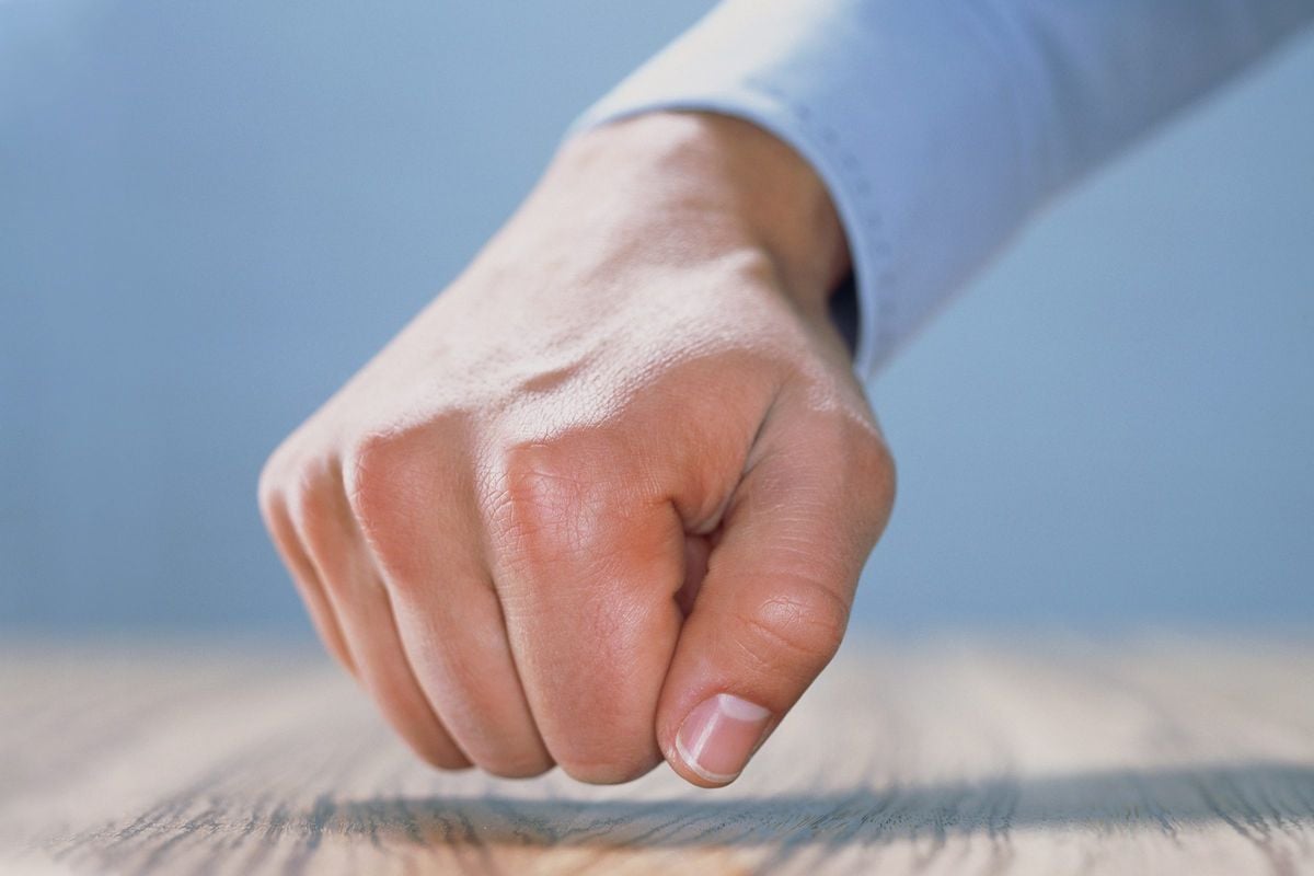 A close-up of a hand with a clenched fist pressing down on a wooden surface, with a light blue background.