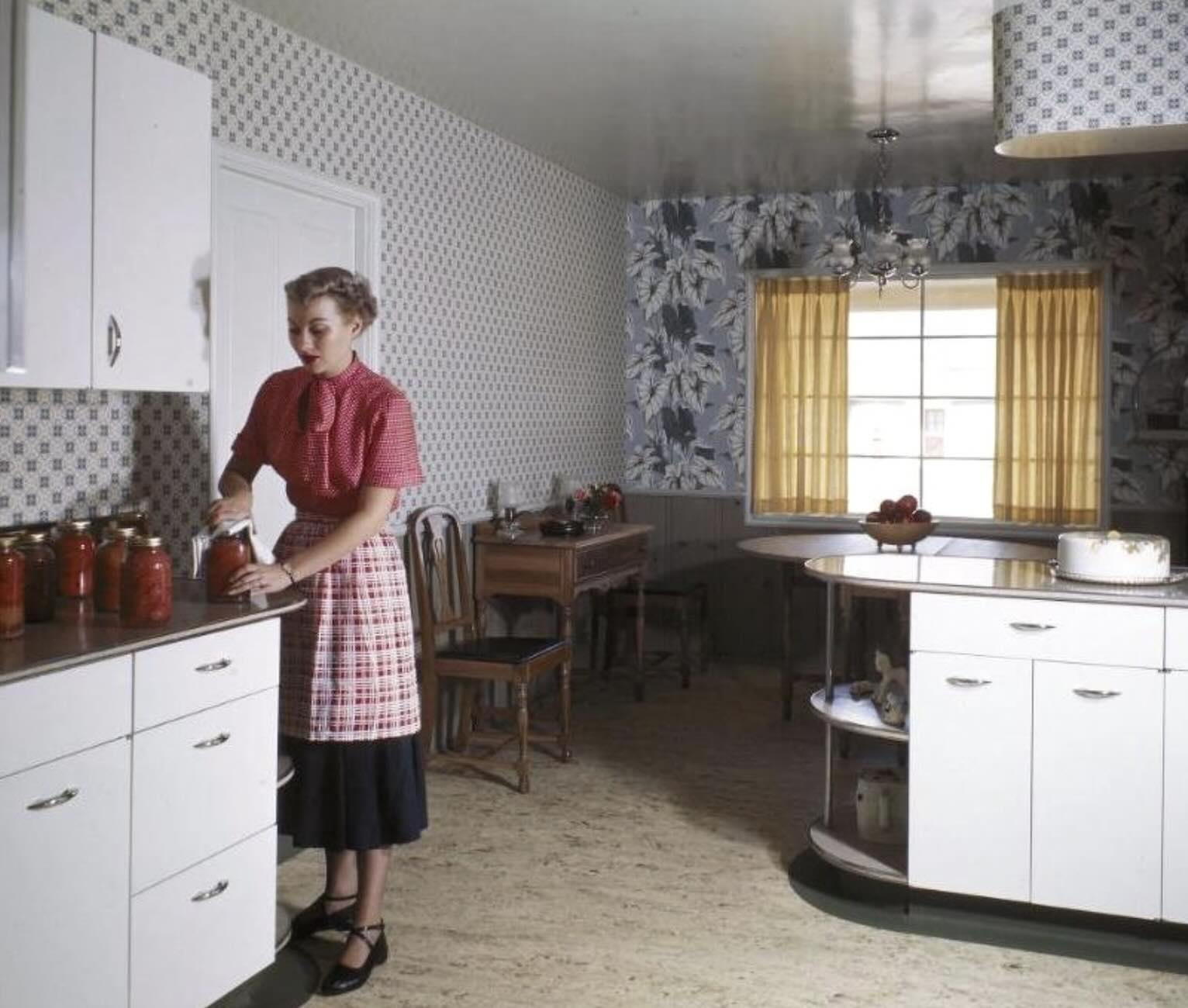 A woman in a vintage kitchen wearing a red blouse and checkered apron stands by a counter with jars. The kitchen has patterned wallpaper, yellow curtains, and white cabinets with a cake on the counter.