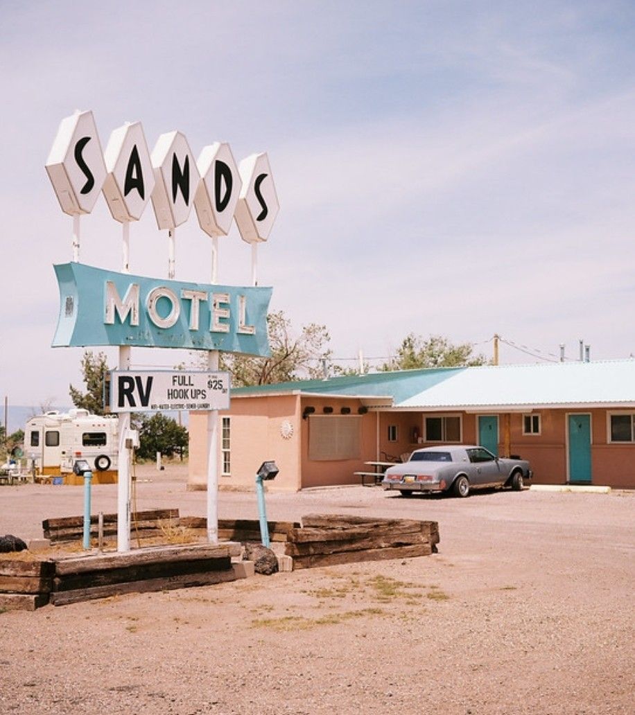 A low-angle shot of the vintage "Sands Motel" sign with diamond-shaped letters. In the background, a peach-colored motel building with blue doors sits in a gravel lot, with a classic gray sedan and a white RV parked nearby under a pale sky.