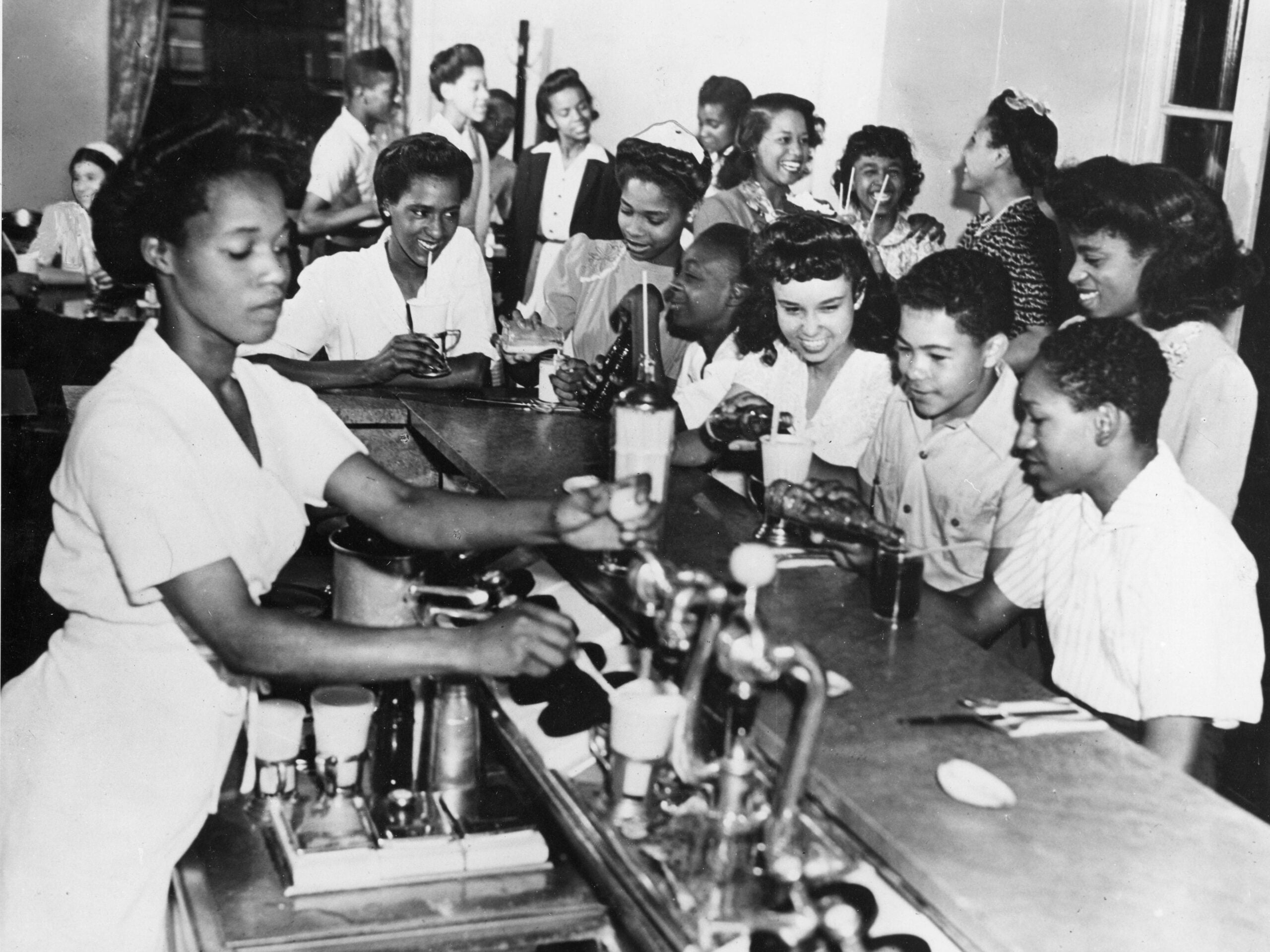 A group of young people sit at a counter in a cafe or soda shop as a woman behind the counter serves drinks; others stand and socialize in the background.
