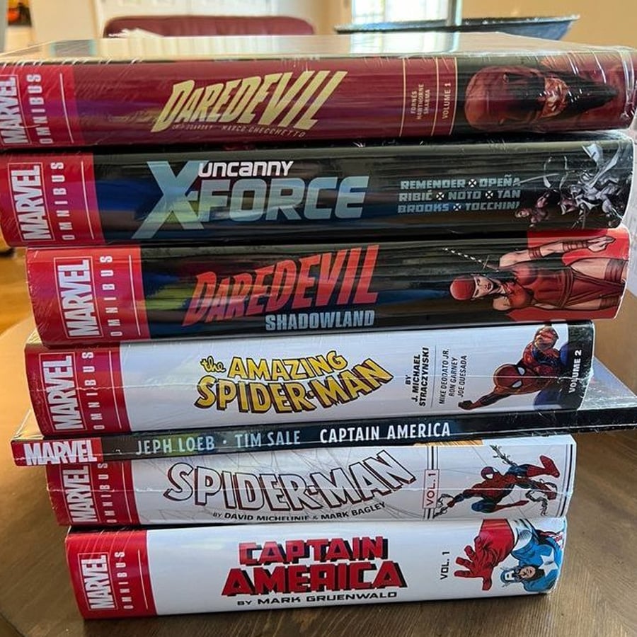A stack of Marvel Omnibus comic books, including Daredevil, Uncanny X-Force, Shadowland, The Amazing Spider-Man, Spider-Man, and Captain America, arranged on a wooden surface.