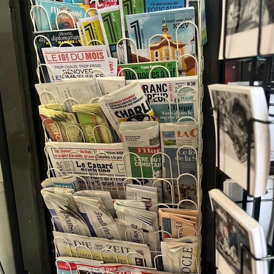 A display rack filled with various French and German newspapers and magazines, including Le Monde diplomatique, Le Figaro, Charlie Hebdo, and Die Zeit, arranged in multiple rows.