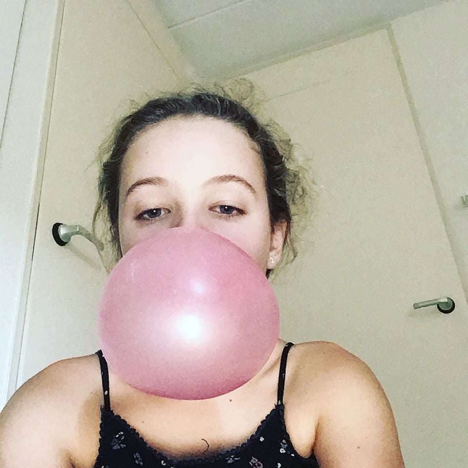 Young person with curly hair blows a large pink bubblegum bubble, nearly covering their nose and mouth. The background shows a white door and walls, and the person is wearing a black spaghetti strap top.
