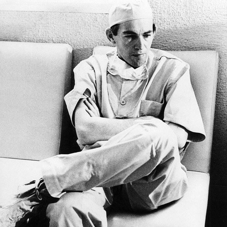 A person in surgical scrubs, cap, and mask sits on a couch with arms crossed, looking thoughtful or tired. The photo is black and white.