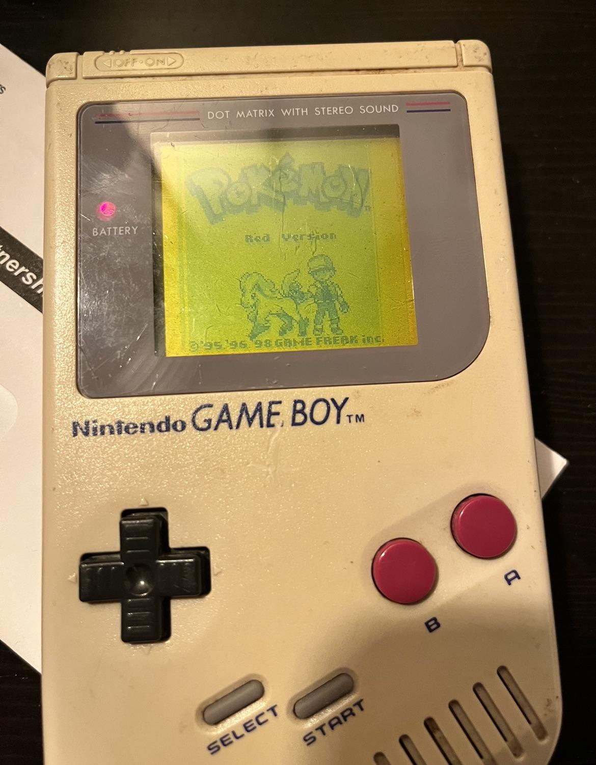 A classic Nintendo Game Boy displaying the title screen for Pokémon Red Version. The screen shows a trainer with a Pokémon. The Game Boy has visible wear and red A and B buttons.
