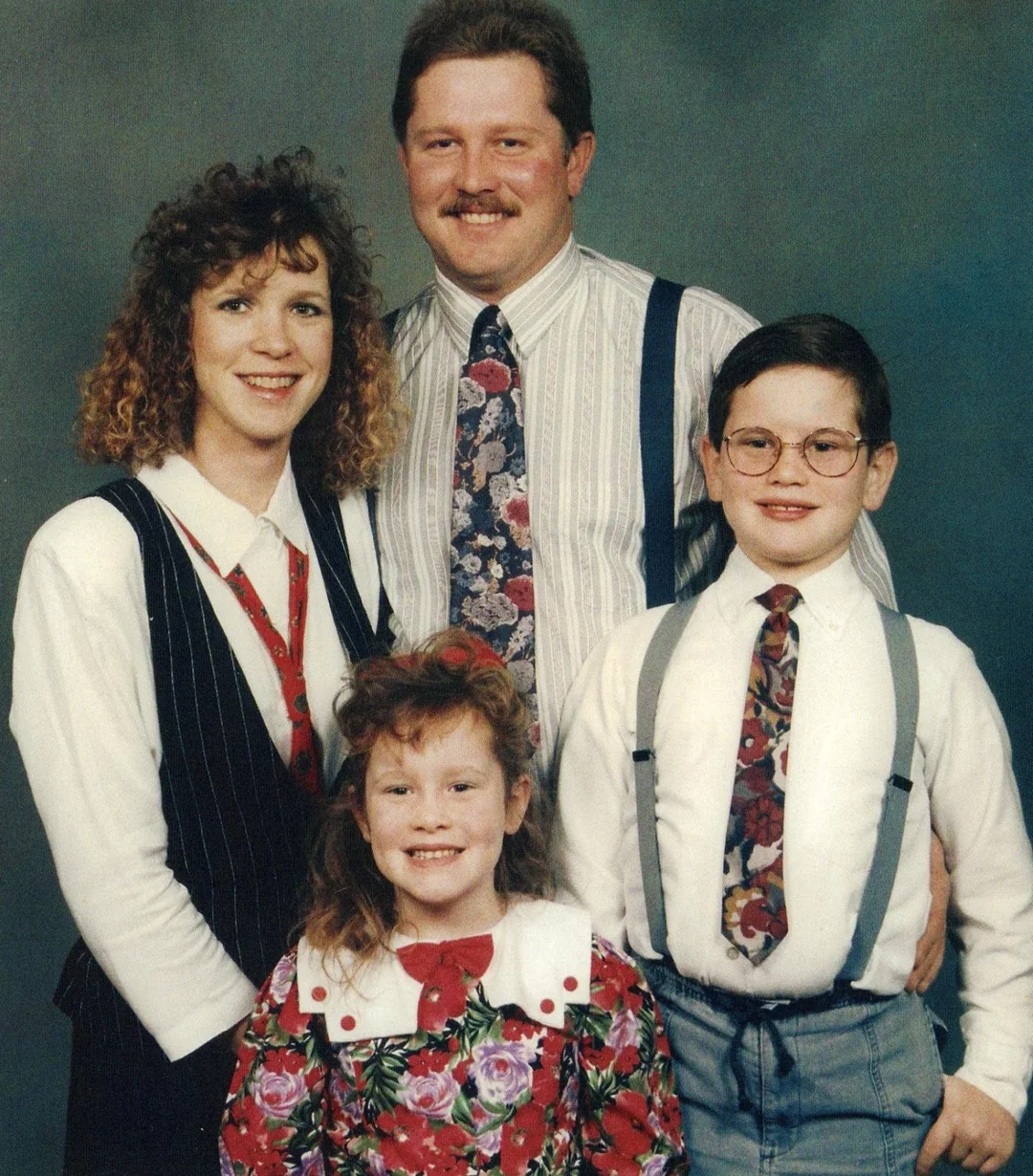 A family of four poses for a portrait in formal attire. The adults stand behind two children; everyone is smiling. The boy wears glasses and suspenders, and the girl in front has curly hair and a floral dress.