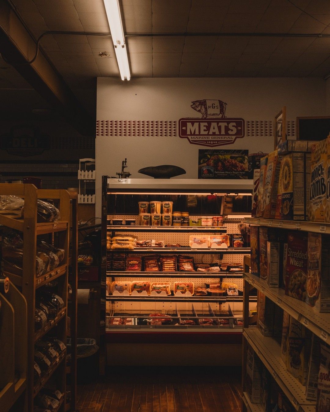 A dimly lit grocery store interior showing shelves of cereal on the right and a refrigerated meat section in the center, with a sign above reading "Meats" and various packaged meats displayed inside.