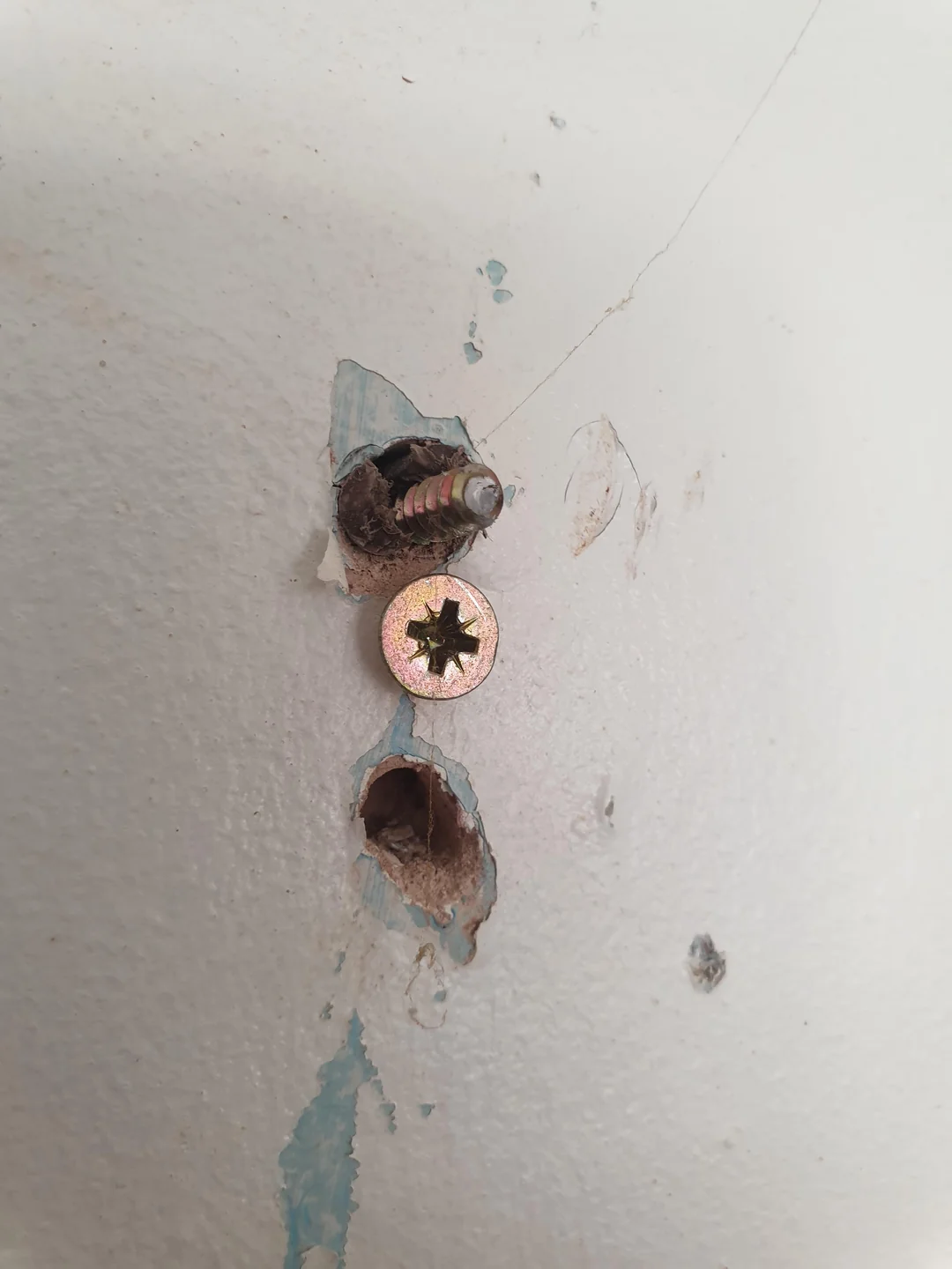 A wall with a damaged area showing two screws, one partially out and at an angle, above a second, fully inserted screw. The paint around the holes is chipped and peeling, with some blue stains below the damage.