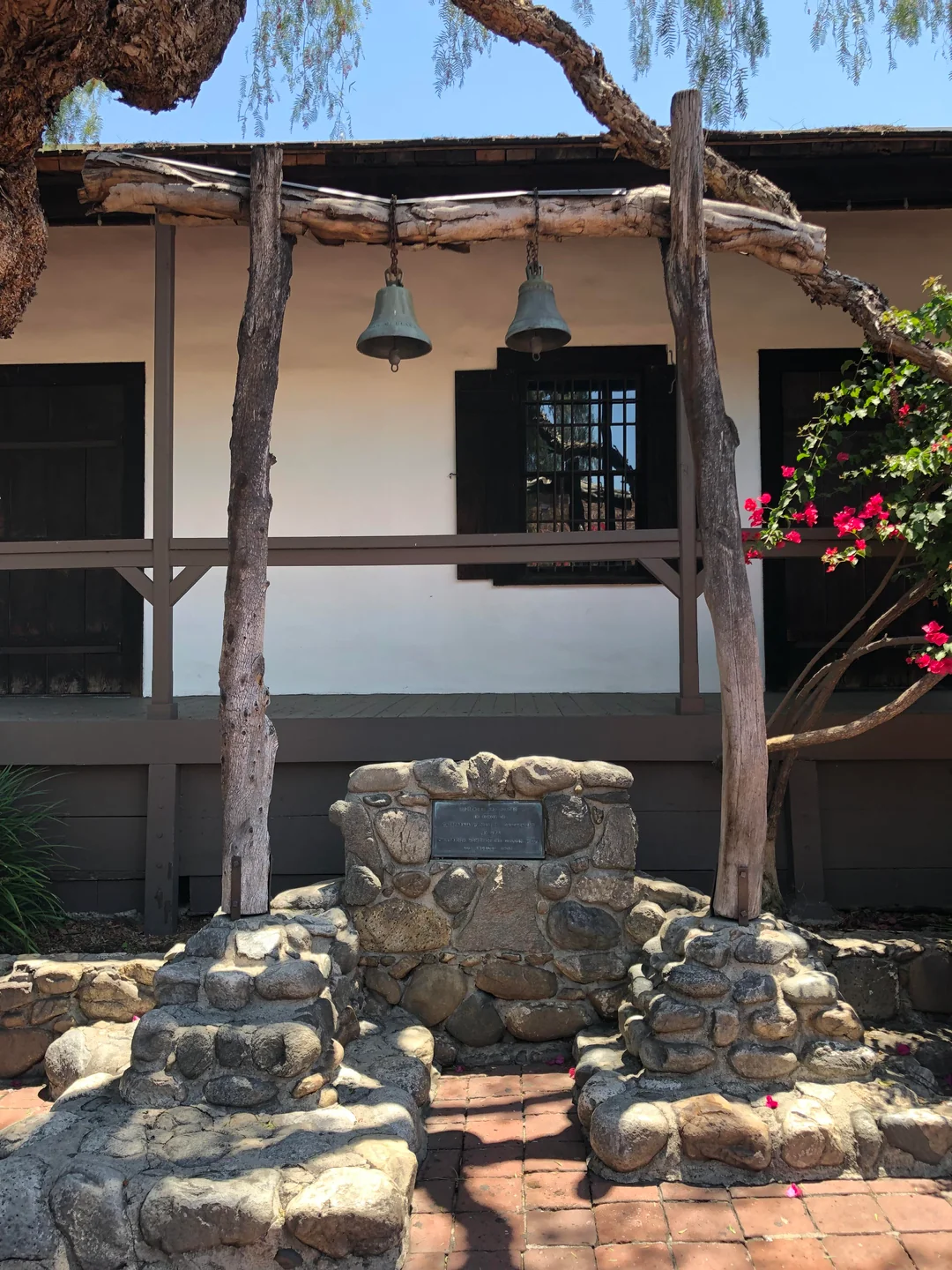 Two large bells hang between rustic wooden posts above a stone monument and plaque, in front of an adobe-style building with wooden porch railings and blooming pink flowers to the right.