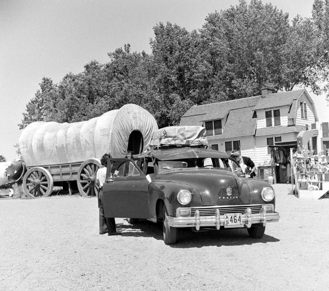 A vintage car with open doors and luggage on the roof is parked in front of a covered wagon and a two-story house. Trees and people are visible in the background on a sunny day.