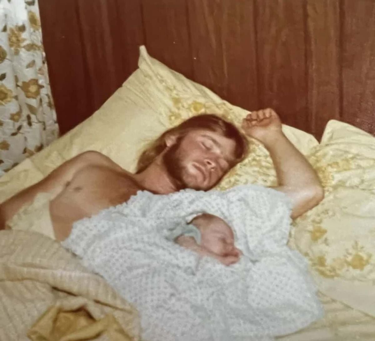 A man with long hair and a beard is sleeping on a bed, cuddled up next to a newborn baby under a blanket, surrounded by pale yellow pillows and bedding with a floral curtain in the background.