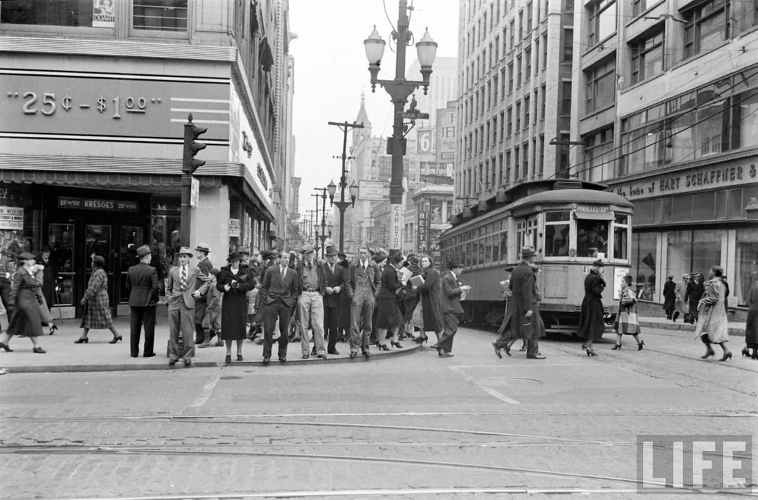 Black and white photo of a busy city street in the 1930s or 1940s, with people crossing the street in front of a streetcar and tall buildings, including shops with large display windows.