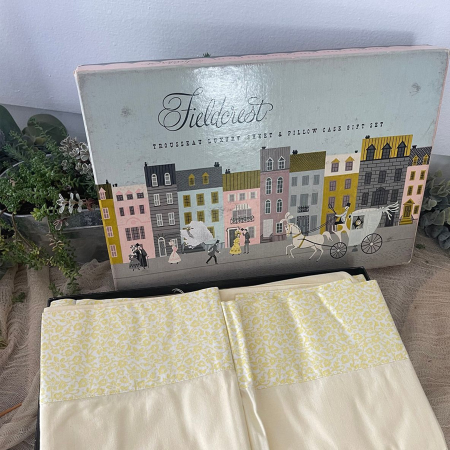 Fieldcrest sheet and pillowcase gift set box with an illustrated street scene, shown behind two folded, cream-colored linens with yellow floral borders, placed on a table with greenery beside them.