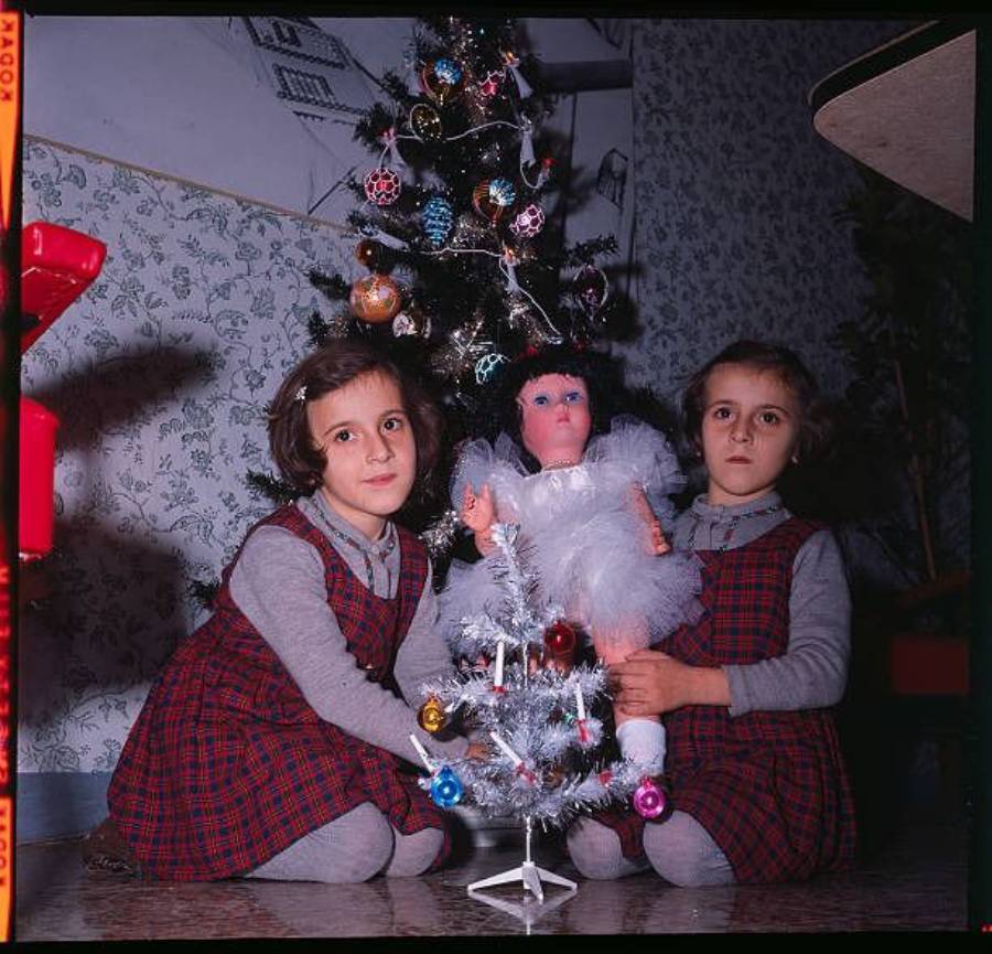 Two young girls in matching plaid dresses sit by a decorated Christmas tree, holding a large doll in white. A small white Christmas tree with ornaments sits in front of them. The scene appears festive and indoors.