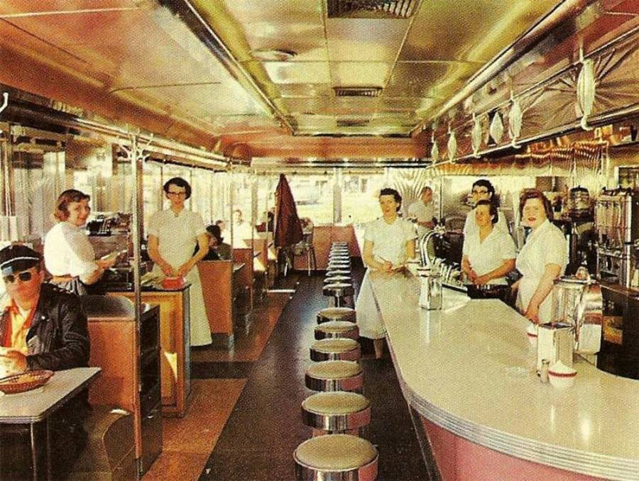 Vintage diner scene with several waitresses behind the counter, a man wearing sunglasses and a cap sits at the counter, and other patrons sit in booths. The décor features chrome details and checkered flooring.