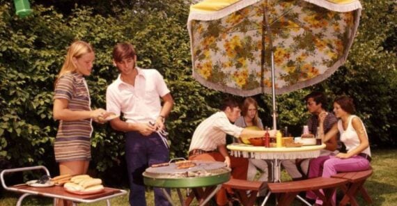 Six people enjoying a backyard barbecue, with two grilling food and four sitting at a table under a floral umbrella. The scene is set on green grass with hedges and trees in the background.