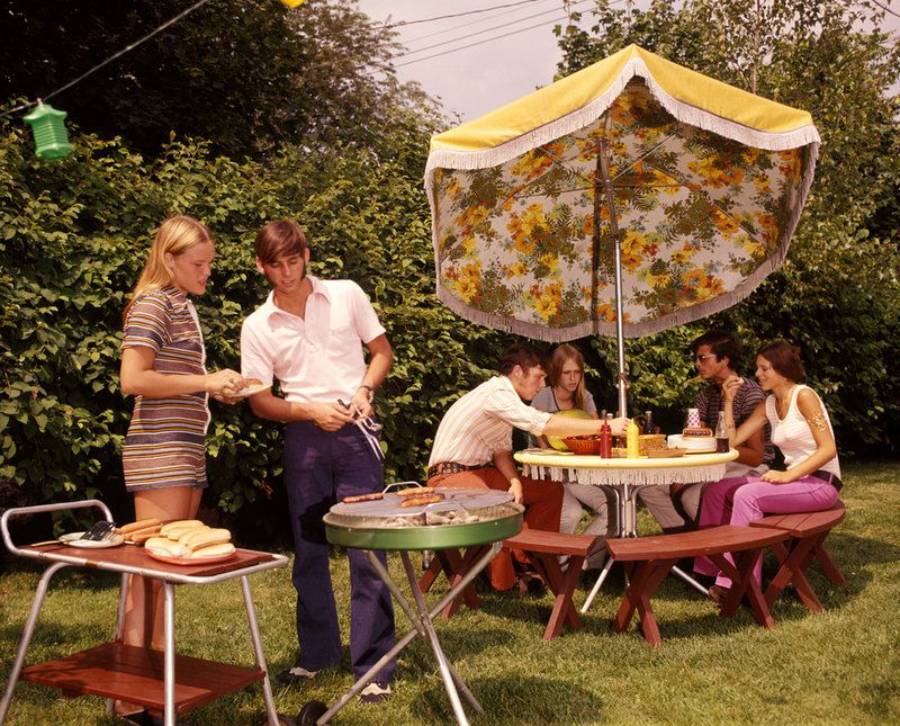 Six people enjoy a backyard barbecue. Two stand by a grill cooking food, while four sit at a table under a floral umbrella, surrounded by green bushes and trees on a sunny day.