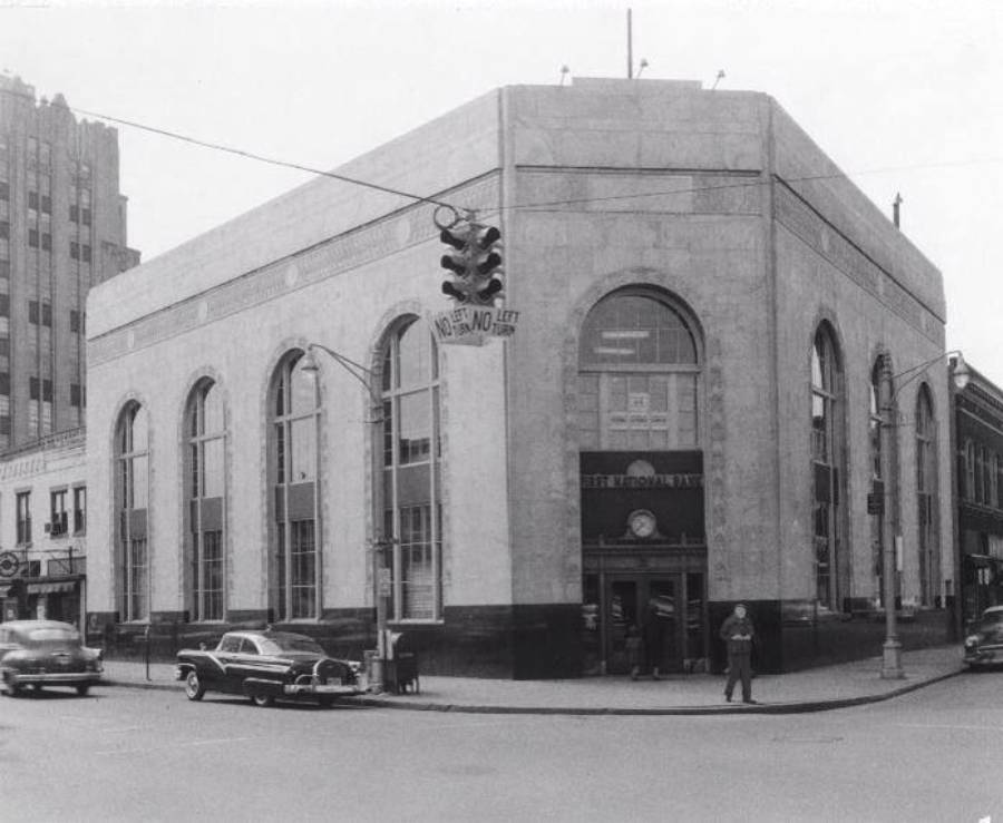 A man stands outside a large, two-story stone bank building with tall arched windows. Vintage cars are parked nearby on the street. A traffic light hangs above the intersection.