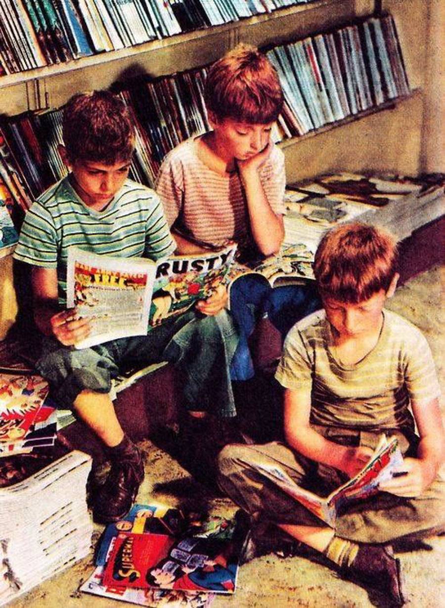 Three young boys sit on the floor and a bench, reading comic books, surrounded by shelves and stacks of more comics in a brightly lit room.