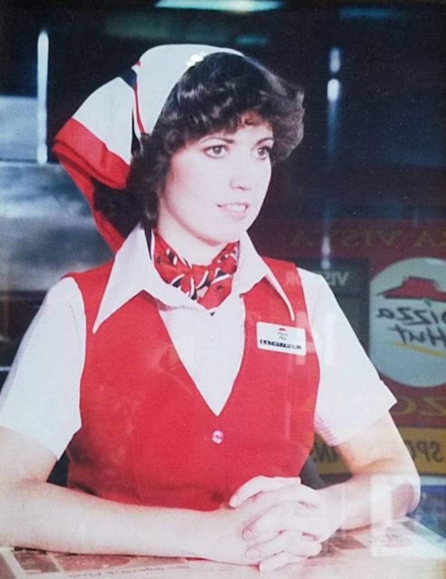 A woman in a red and white uniform with a matching scarf and hat sits at a counter with hands folded. She wears a name tag and has short, curly hair. The background includes Pizza Hut signage.