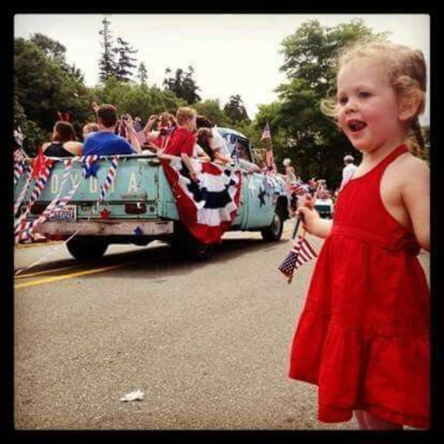 A young girl in a red dress holds a small American flag and smiles near a parade. Behind her, people ride a blue truck decorated with flags and patriotic bunting. Trees and more parade participants are visible in the background.