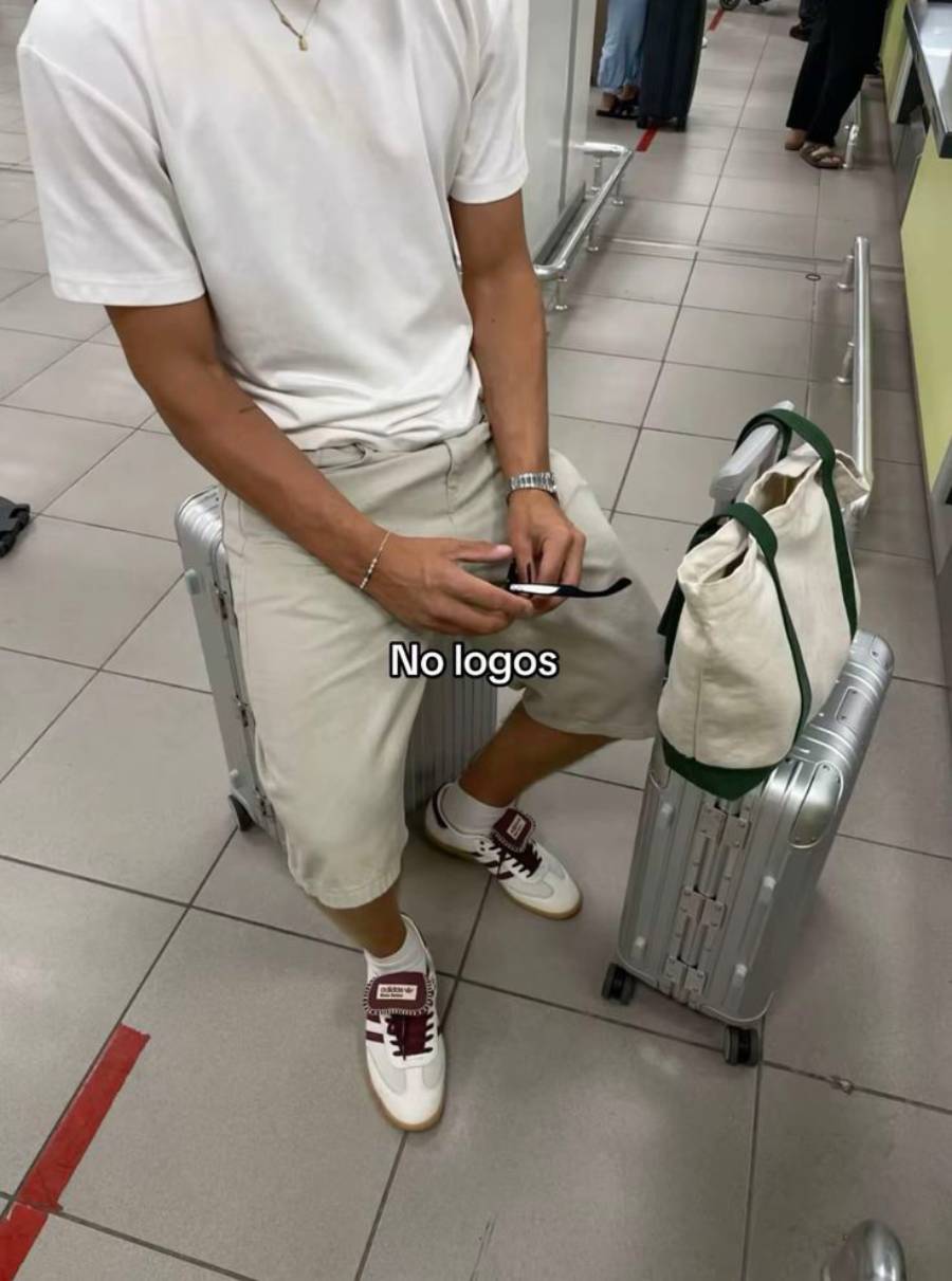 A person in a white t-shirt and light shorts sits on a suitcase, holding a phone with another suitcase and a tote bag beside them. The text "No logos" is overlaid on the image.