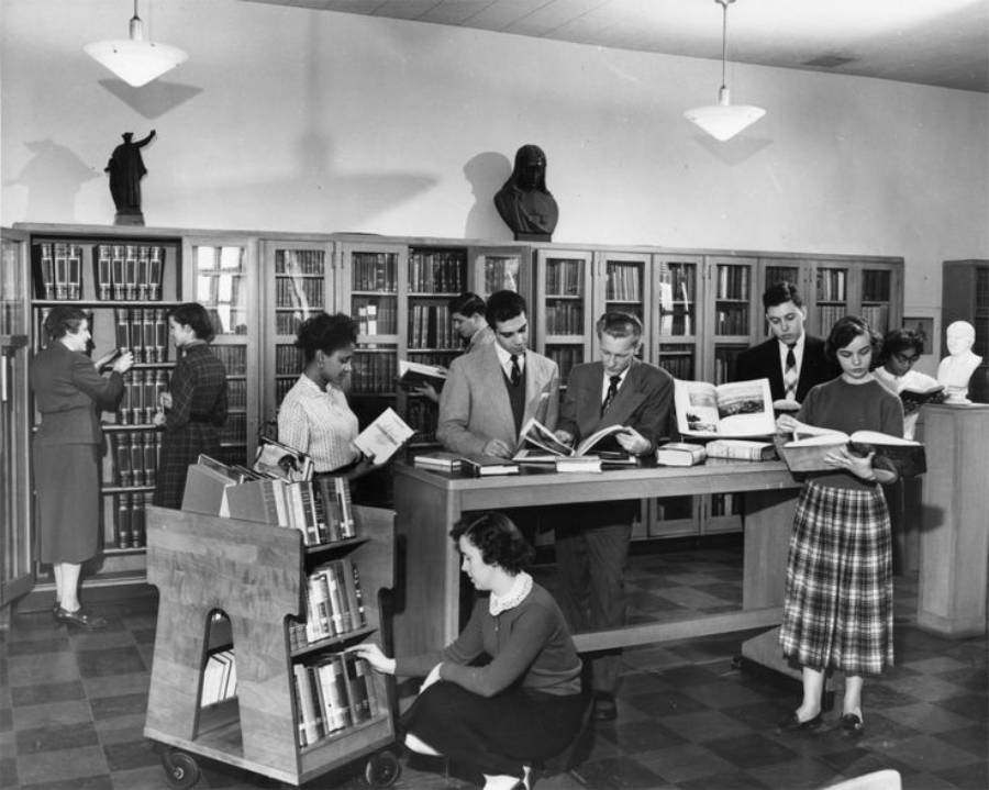 Black and white photo of people browsing and reading books in a library. Some stand at a table, others select books from shelves or a cart. Busts and statues are visible in the background.