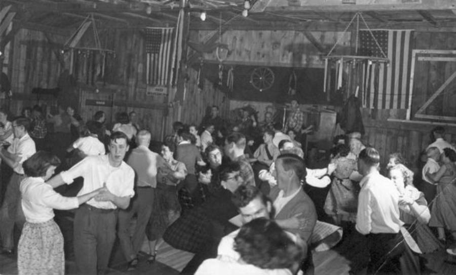 Black and white photo of people dancing in pairs inside a rustic hall, with American flags and wooden beams visible in the background. The room appears lively and crowded.