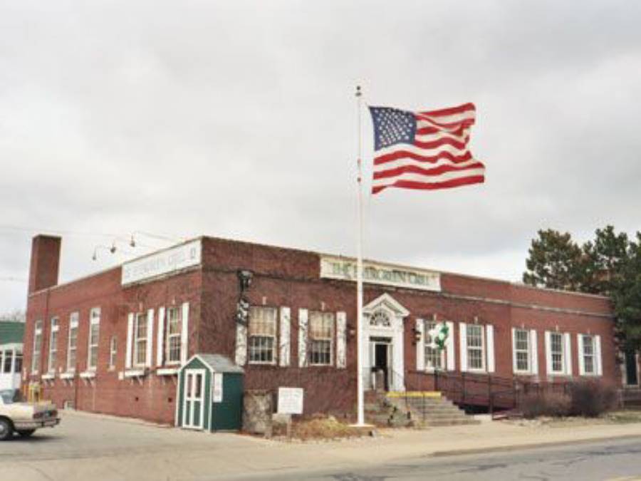 A large American flag waves in front of a one-story brick building with white trim and an arched entrance on a cloudy day. A small green structure is near the sidewalk. A car is parked on the street.