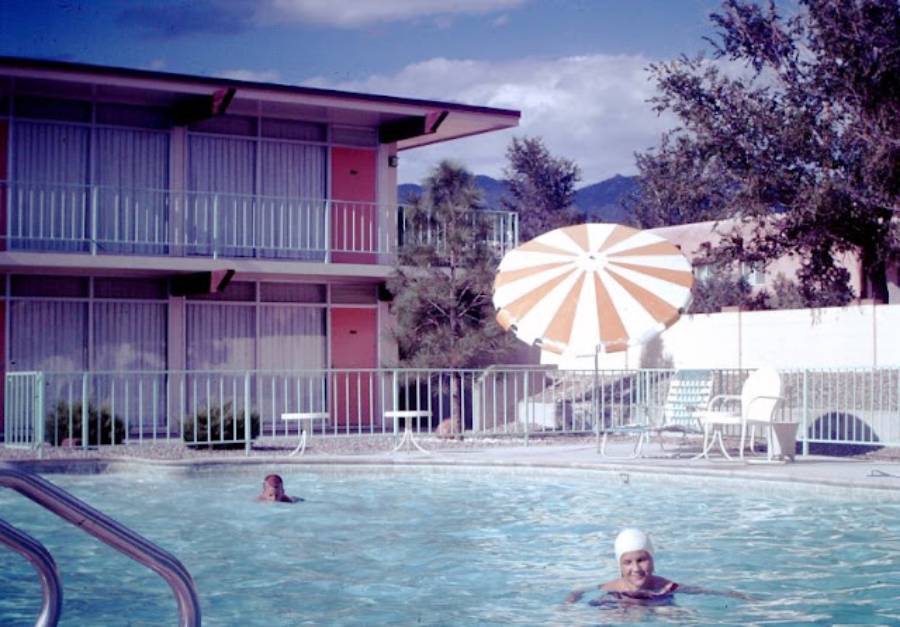 Two people swim in a clear outdoor hotel pool. There are empty white chairs, a large orange-and-white umbrella, and a modern two-story building with glass windows in the background. Trees and mountains are visible.