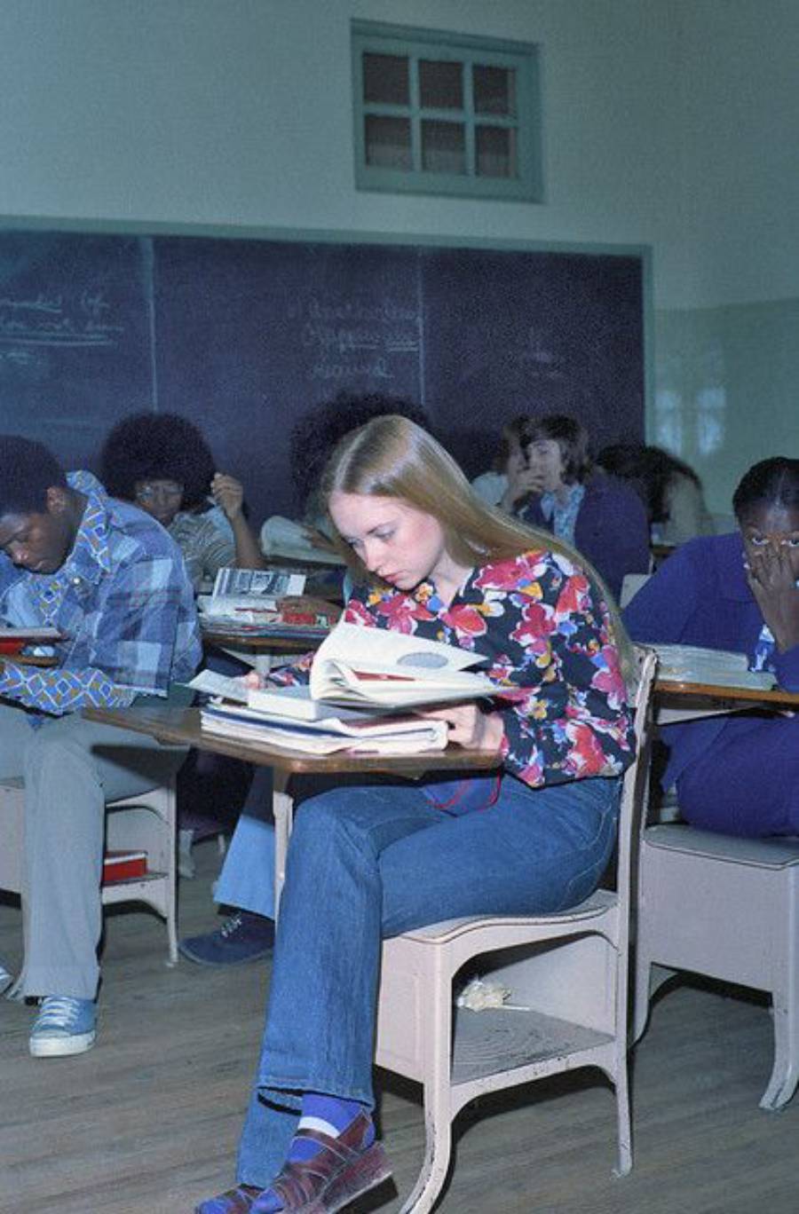 A young woman with long blonde hair, wearing a floral shirt and jeans, sits at a school desk reading a book. Other students are seated around her in a classroom with a blackboard in the background.