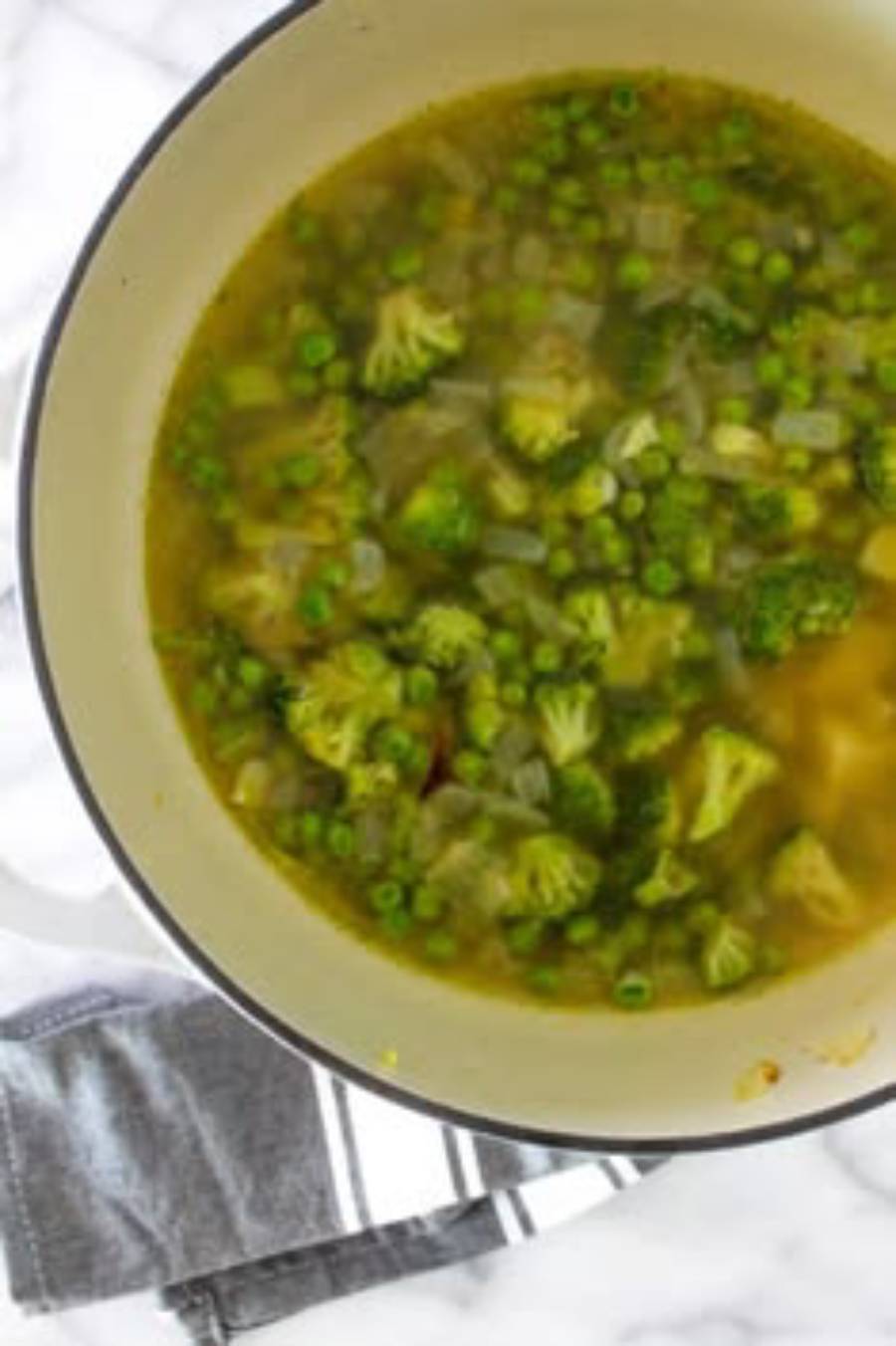 A pot of soup with broccoli florets, green peas, and chopped onions sits on a white surface next to a folded gray cloth and a striped towel.