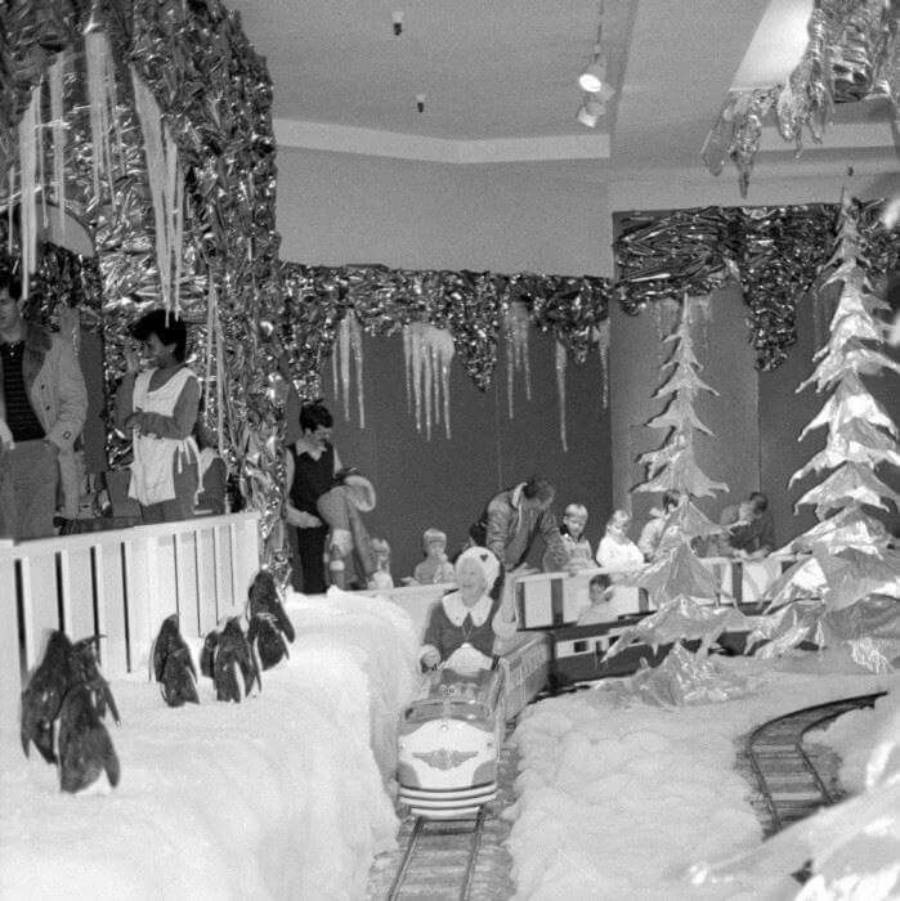 Black and white photo of a wintry indoor train ride with artificial snow, penguin figures, shiny icicles, and people watching children on a small train track.