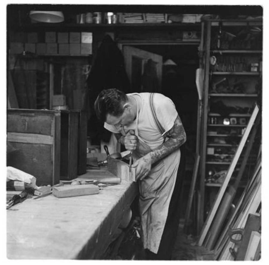 A man wearing an apron and face mask uses a chisel on a piece of wood at a workbench in a cluttered workshop with shelves, boxes, and tools in the background.