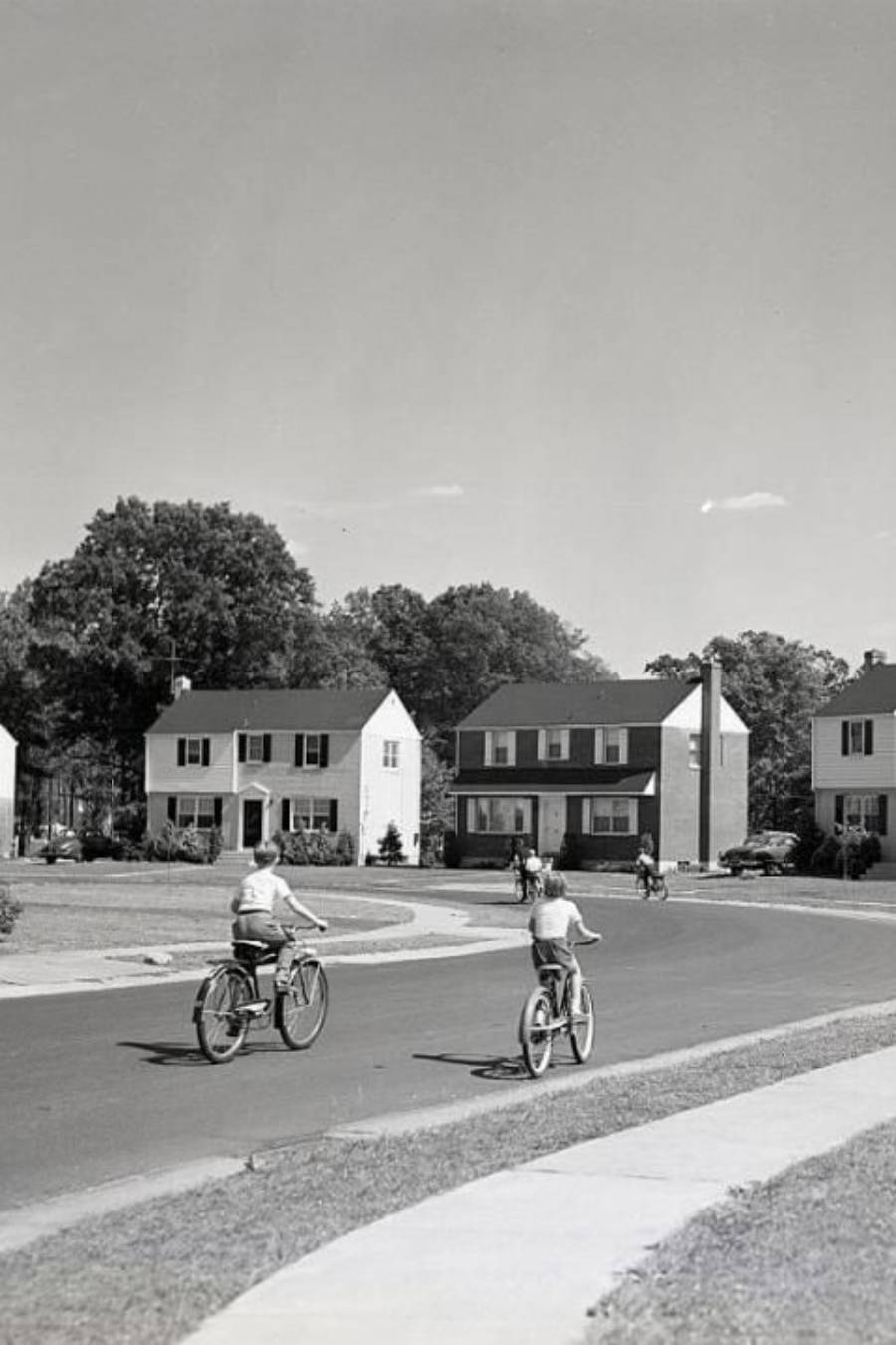 Black-and-white photo of children riding bicycles on a quiet suburban street, with two-story houses and trees in the background, under a clear sky.