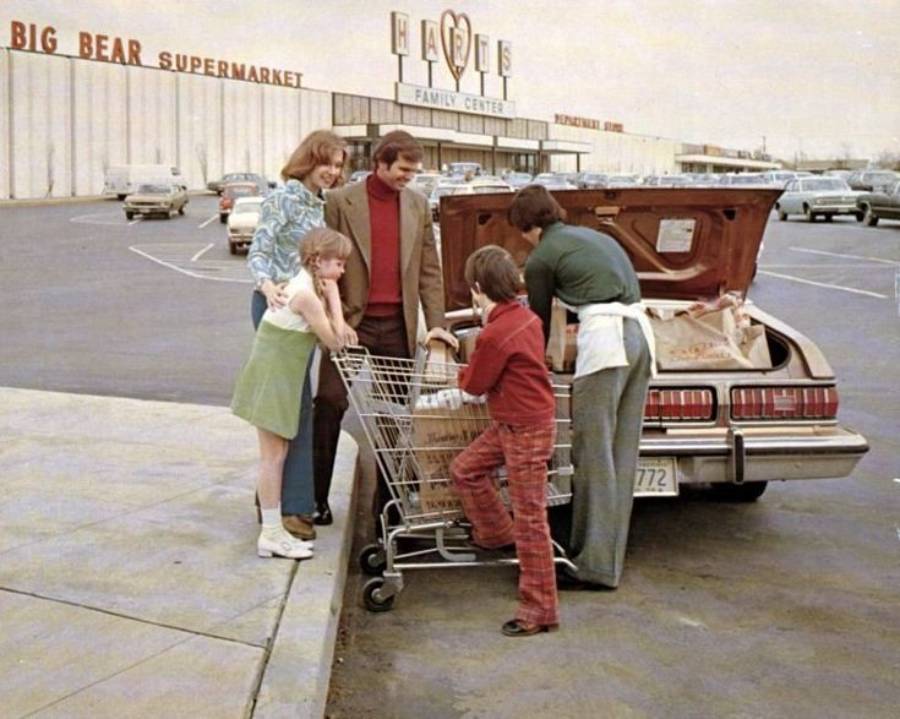 A family stands by their car in a supermarket parking lot, loading groceries from a cart into the trunk. The store sign reads "Big Bear Supermarket" and "Harts Family Center.
