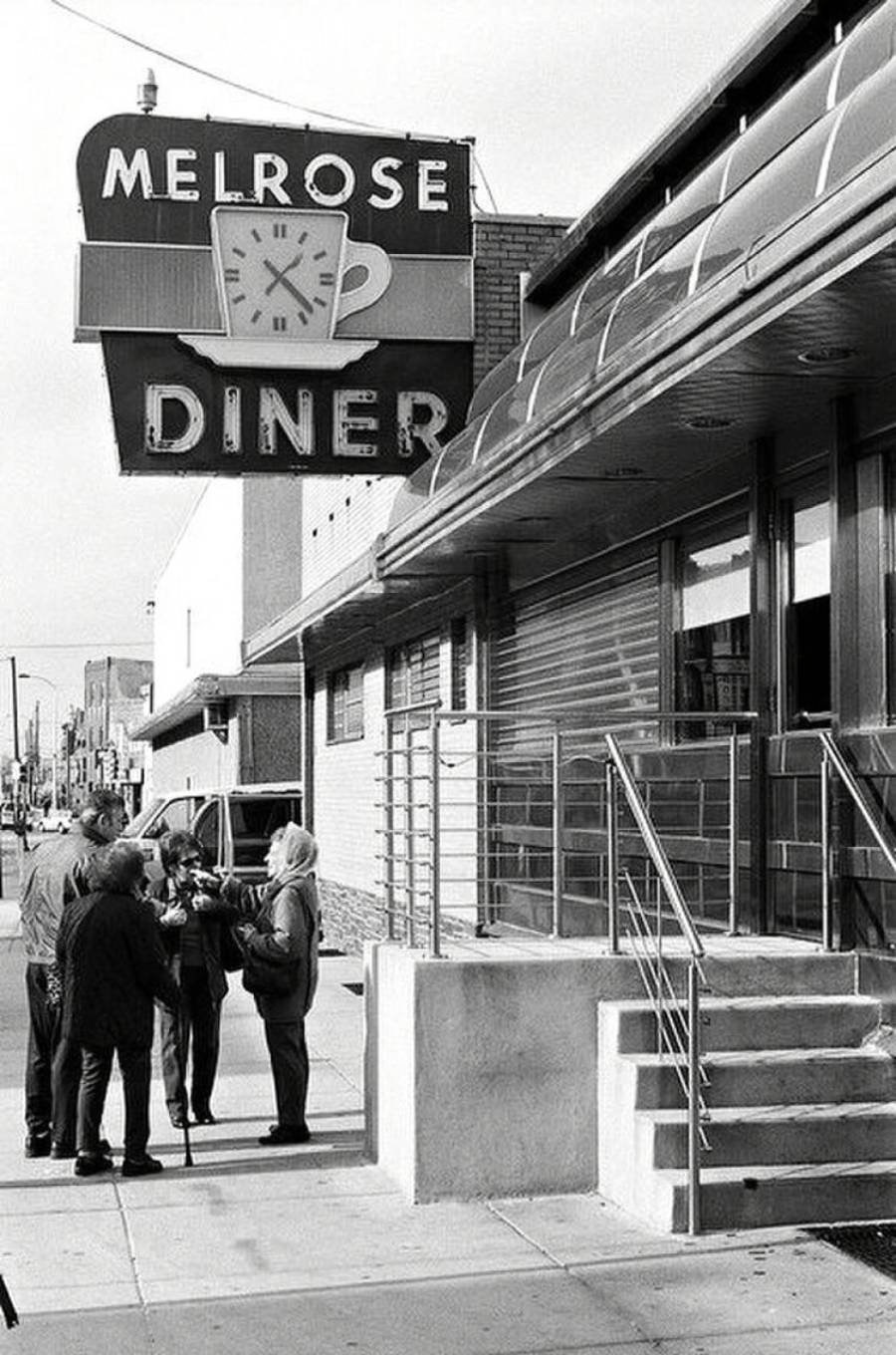 Black and white photo of several people talking outside the Melrose Diner, which has a large sign featuring a coffee cup with a clock on it. The building has steps and a metal awning.