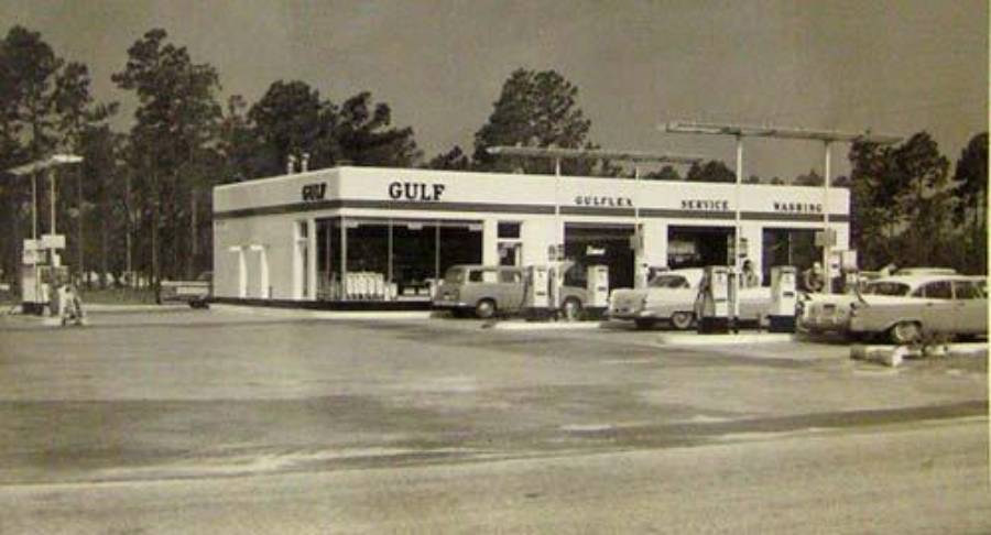A vintage black-and-white photo of a Gulf gas station with several cars parked at the pumps. The building displays signs for Gulf, service, and washing. Trees are visible in the background.