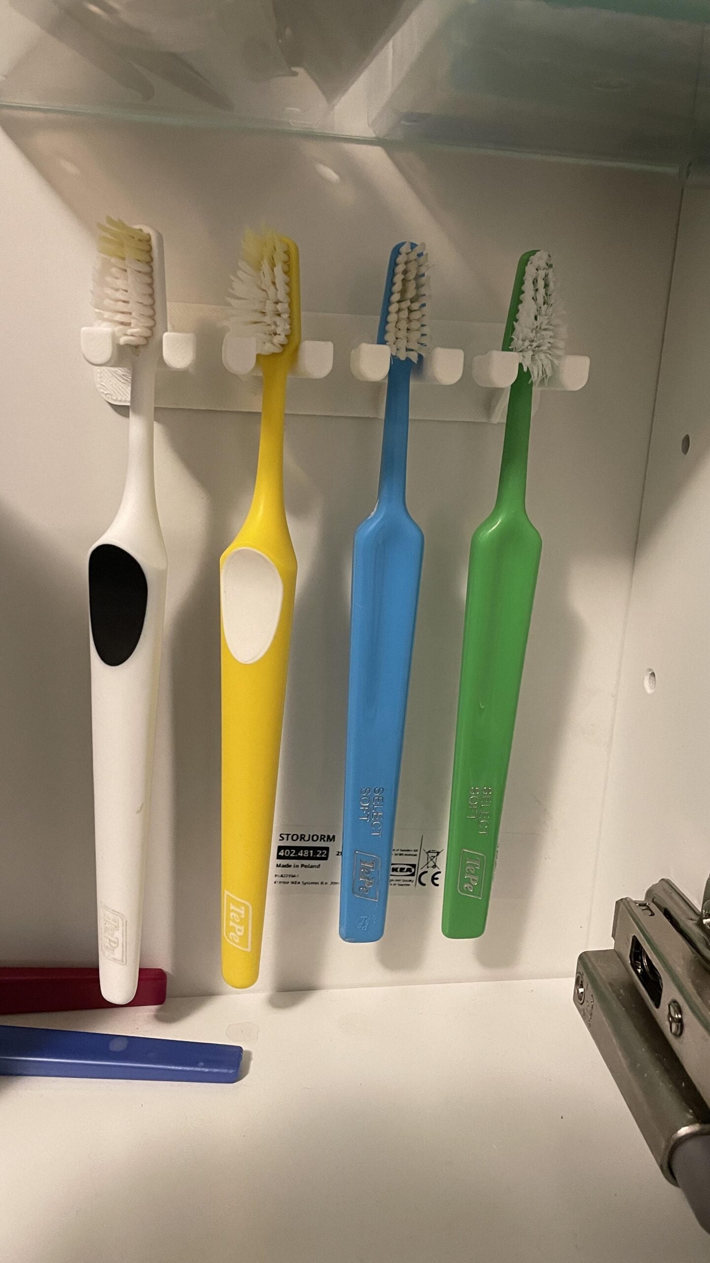 Four toothbrushes—white, yellow, blue, and green—hang neatly on a holder inside a bathroom cabinet. A metal object and a blue item are visible at the bottom right and left corners.