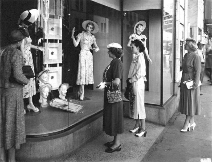Four women stand on a sidewalk, looking at mannequins dressed in 1940s-style clothing displayed in a store window. The mannequins wear hats and dresses; two women hold handbags.