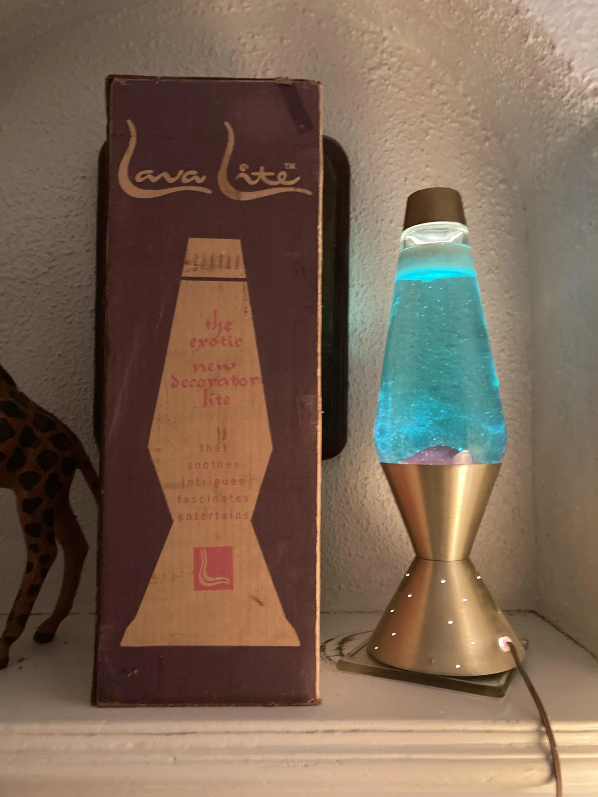 A blue lava lamp glows beside its vintage box, which reads "Lava Lite: the exotic new decorator kit." A giraffe statue is partially visible on the left. The scene is set on a white shelf.