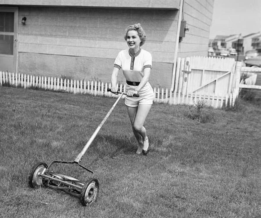 A smiling woman in shorts and a t-shirt pushes a manual reel lawn mower on a grassy lawn, with a white picket fence and a house in the background.