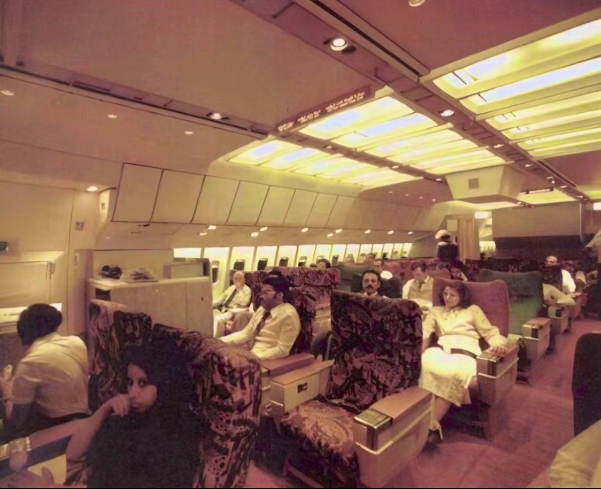 Passengers sit in a spacious, retro airplane cabin with patterned seats, soft yellow lighting, and large windows. Some people are relaxing, while others are engaged in conversation or looking around.