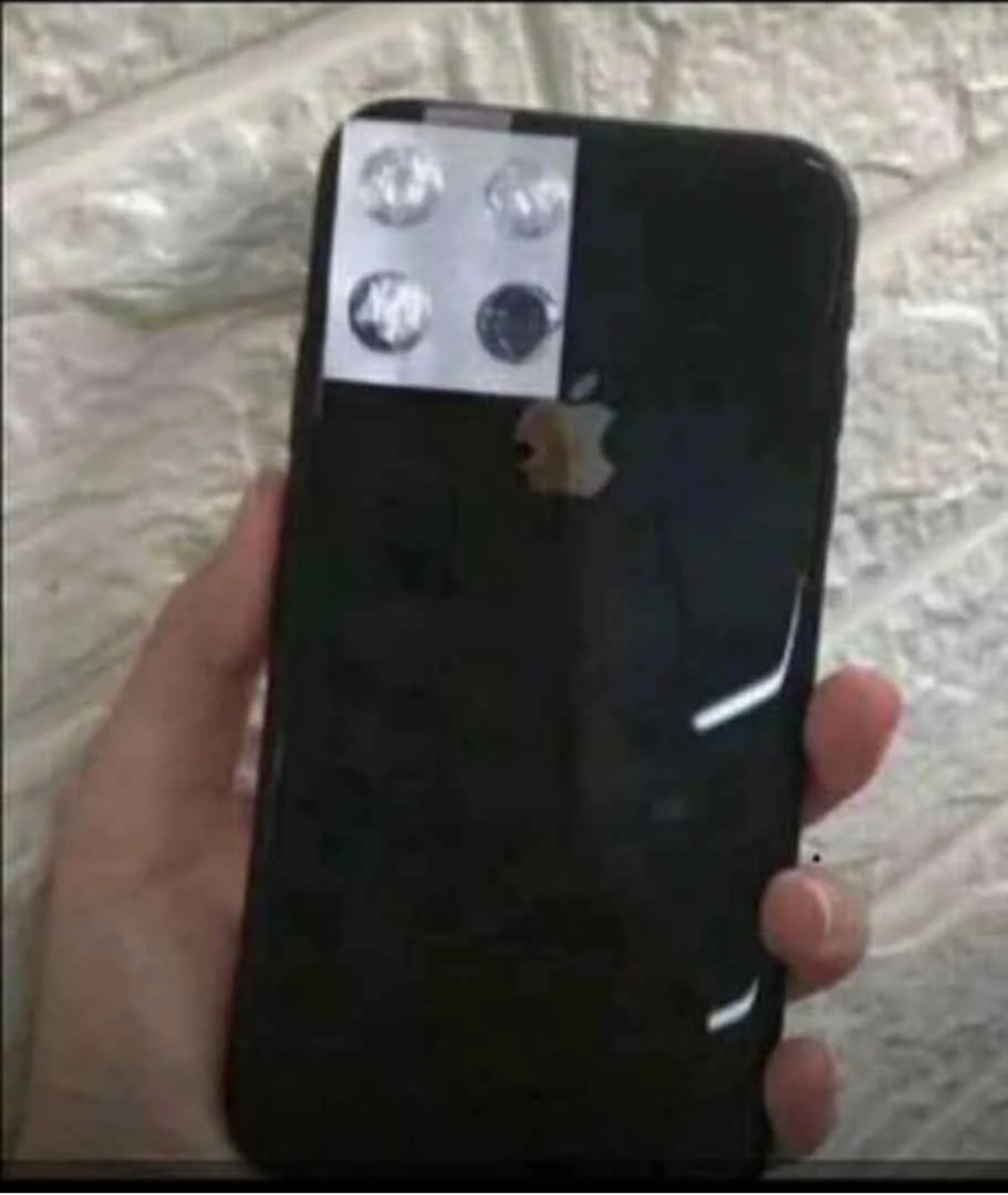 A hand holds a black smartphone with a fake camera setup made from foil and paper, mimicking multiple rear cameras, against a textured white wall.