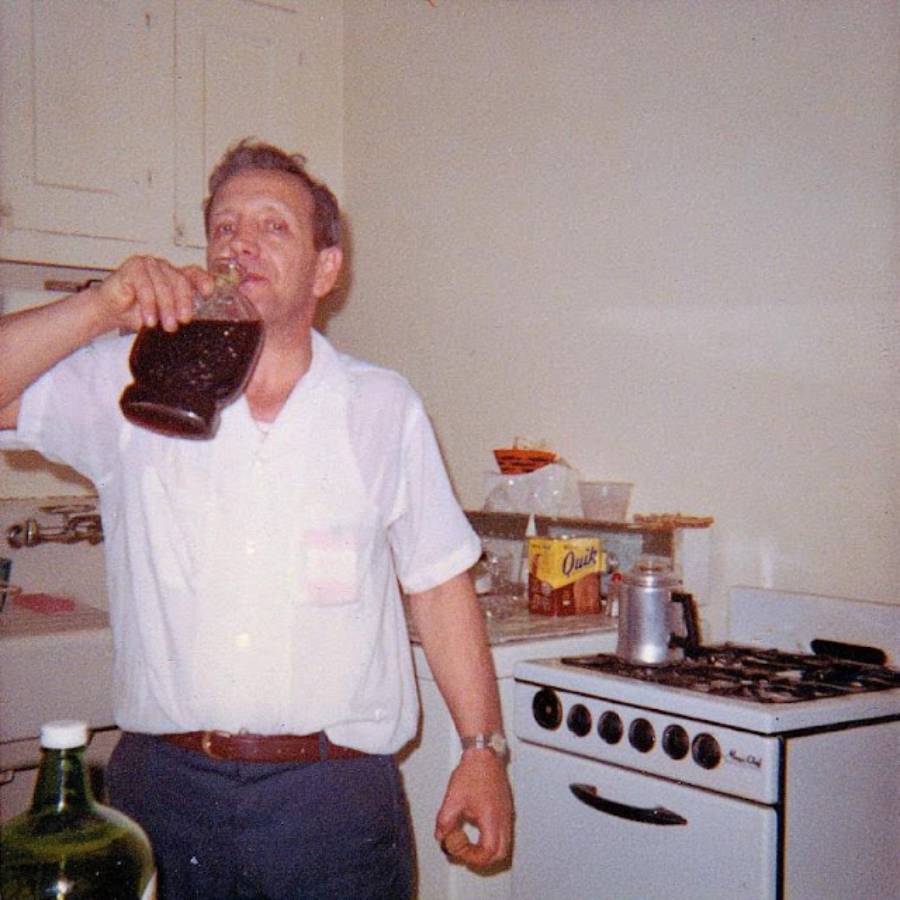 A man in a white shirt drinks from a large glass jug in a kitchen. A box of Quik and other items are on the counter near a stove.