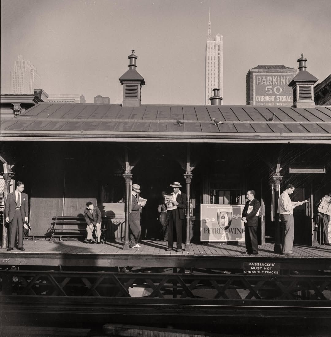 Commuters stand and sit on a vintage train platform, reading newspapers and waiting. Buildings and a parking sign are visible in the background.